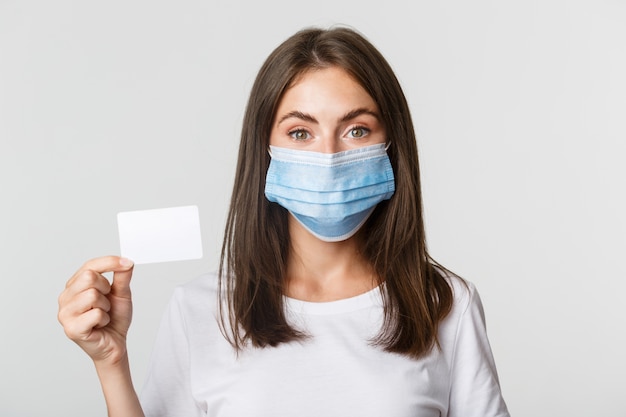 Covid-19, health and social distancing concept. Close-up of pretty brunette girl in medical mask, showing credit card.