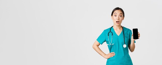 Covid-19、医療従事者とオンライン医療の概念。興奮して驚いたアジアの女性看護師、医師は携帯電話の画面、インターネット相談アプリ、白い背景を表示しながら驚いて見えます。