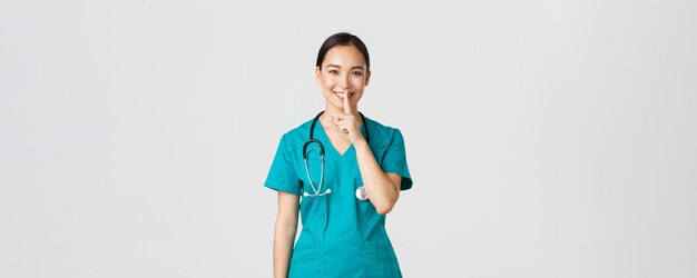 Covid-19, healthcare workers, pandemic concept. Cheerful, smiling female asian nurse in scrubs having secret, making surprise, showing shush gesture, shhh keep quiet, standing white background.