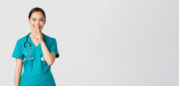 Covid-19, медицинские работники, концепция пандемии. Веселая, улыбающаяся азиатская медсестра в халате, имеющая секрет, удивляющая, показывающая шш-жест, тсс-с, молчи, стоя на белом фоне.