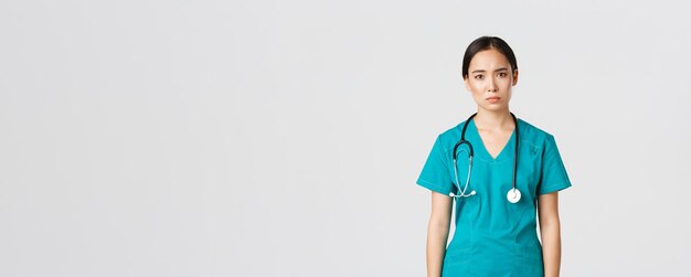 Covid-19, healthcare workers, pandemic concept. Exhausted young asian female nurse, doctor looking tired after shift in hospital, looking sad with fatigue, standing white background in scrubs.