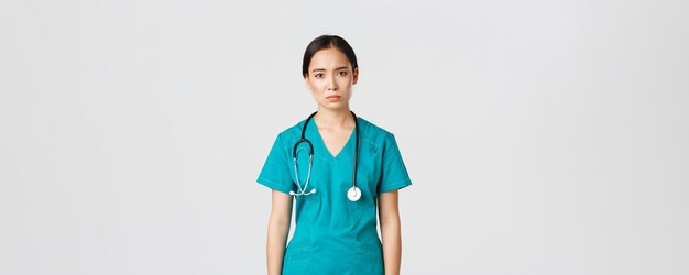 Covid-19, healthcare workers, pandemic concept. Exhausted young asian female nurse, doctor looking tired after shift in hospital, looking sad with fatigue, standing white background in scrubs.