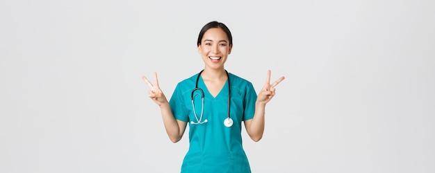 Covid-19、医療従事者、パンデミックの概念。幸せなアジアの女性医師、平和カワイイジェスチャーを示して笑顔でスクラブの看護師、病院での仕事中に前向きなまま、白い背景