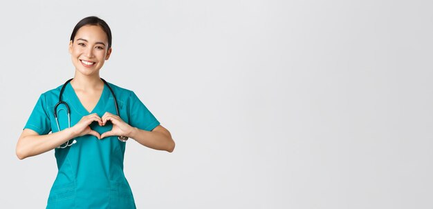 Covid-19, медицинские работники, концепция пандемии. Прекрасный заботливый азиатский врач, медсестра в скрабах, показывая жест сердца и улыбаясь, заботясь о пациентах с любовью, на белом фоне.
