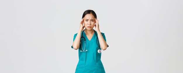 Covid-19, healthcare workers, pandemic concept. Overworked and exhausted asian female doctor, physician feeling sick, wear scrubs, touching head, complaining on headache or high fever