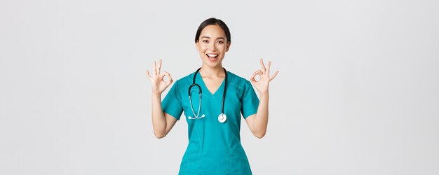 Covid-19, healthcare workers, pandemic concept. Professional confident asian female doctor, intern in scrubs assure patients everything good, showing okay gesture satisfied, smiling pleased