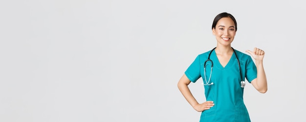 코비드-19, 의료 종사자, 전염병 개념. 전문적인 자신감 있는 아시아 여성 의사, 수술복을 입은 간호사가 자신을 가리키며 웃고 있으며, 그녀가 무엇을 하는지 알고 있음을 보장합니다, 흰색 배경