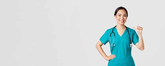 Covid-19, медицинские работники, концепция пандемии. Профессиональная уверенная женщина-врач, медсестра в халате, указывающая на себя и улыбающаяся, гарантирует, что она знает, что делать, белый фон