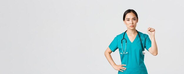 Covid-19, healthcare workers, pandemic concept. Serious-looking confident serious asian female doctor showing support to fellow collegues during coronavirus, raising fist in unity gesture