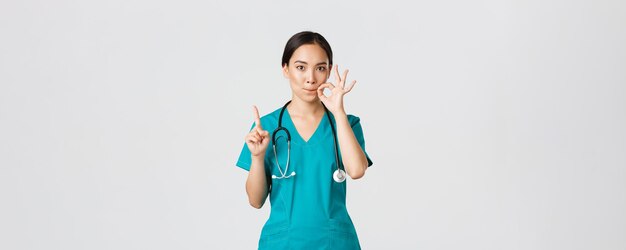 Covid-19, healthcare workers, pandemic concept. Serious-looking worried asian female nurse, physician asking keep secret, shaking finger and showing mouth seal, zipping lips gesture