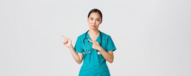 Covid-19, healthcare workers, pandemic concept. Skeptical and doubtful asian female nurse in scrubs pointing upper left corner and grimacing unamused, dont like product, being hesitant