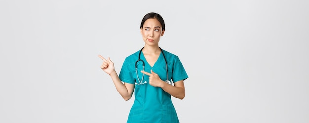 Covid-19, healthcare workers, pandemic concept. Skeptical and doubtful pretty asian doctor, nurse in scrubs smirk, pointing upper left corner, looking with reluctant hesitant face, white background.