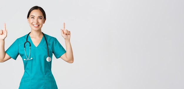 Covid-19, healthcare workers, pandemic concept. Smiling beautiful asian female nurse, doctor in scrubs pointing and looking up with satisfied grin, happy to show awesome promo offer, white background.