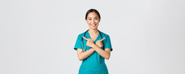 Covid-19, healthcare workers, pandemic concept. Smiling beautiful asian nurse, intern or doctor in scrubs pointing sideways and looking at camera, showing variants, two choices, white background.