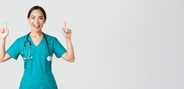 Covid-19, healthcare workers, pandemic concept. Smiling cheerful pretty nurse, female doctor or intern in scrubs pointing fingers up, showing banner, make announcement, demonstrate advertisement.