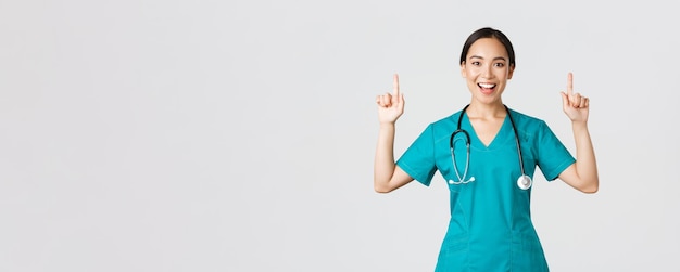 Covid-19, healthcare workers, pandemic concept. Smiling cheerful pretty nurse, female doctor or intern in scrubs pointing fingers up, showing banner, make announcement, demonstrate advertisement