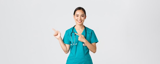 Covid-19, healthcare workers, pandemic concept. Smiling pretty asian doctor, nurse in scrubs and stethoscope, pointing fingers upper left corner, showing online clinic banner, white background.