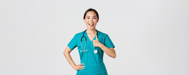 코비드-19, 의료 종사자, 전염병 개념. 웃고 있는 낙관적이고 자신감 있는 아시아 여성 간호사는 모든 것을 통제하고 승인에 엄지손가락을 치켜들고 진료소에서 서비스 품질을 보장합니다