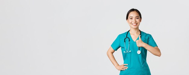 Covid-19、医療従事者、パンデミックの概念。笑顔で明るい、自信を持ってスクラブをしているアジアの女性看護師は、すべてをコントロールし、承認を得て親指を立て、クリニックでのサービスの質を保証します