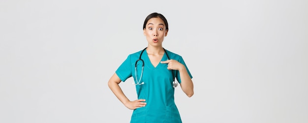 코비드-19, 의료 종사자, 전염병 개념. 놀라고 매복한 아시아 여성 의사, 수술복을 입은 간호사가 자신을 가리키고, 이름이 지정되거나 선택된 흰색 배경에 서 있습니다.
