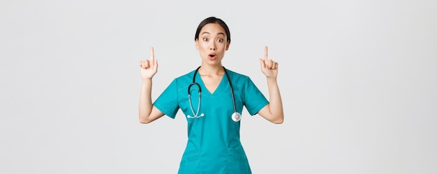 Covid-19, healthcare workers, pandemic concept. Surprised and thrilled asian female nurse, woman doctor in scrubs asking question interesting promo, pointing fingers up, showing clinic advertisement.