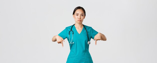 Covid-19, healthcare workers, pandemic concept. Upset and disappointed pouting asian female therapist, nurse in scrubs pointing fingers down, sulking sad, showing something with regret.