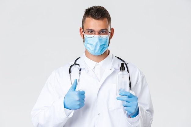 코비드-19, 의료 종사자, 전염병 및 예방 바이러스 개념. 의료용 마스크와 장갑을 끼고 있는 자신 있는 진지한 의사의 클로즈업, 엄지손가락을 위로 올려주는 조언 좋은 손 소독제, 코로나바이러스로부터 보호