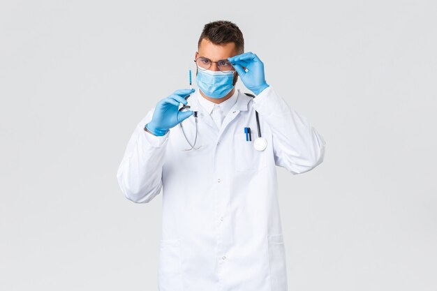 Covid-19、医療従事者、パンデミックおよびウイルス予防の概念。白衣、眼鏡、医療用マスクを身に着けた真面目な専門医は、コロナウイルスワクチンの注射器に興味を持っています。