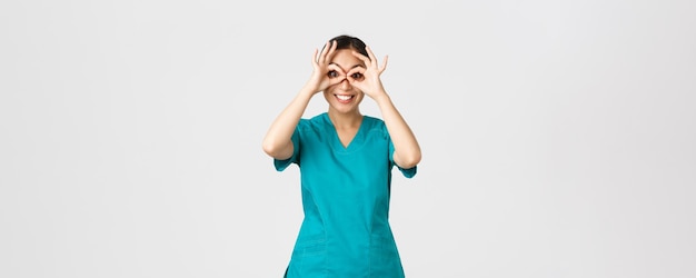 Covid-19、医療従事者とウイルスの概念を防ぐ。面白がって幸せでかわいいアジアの女性医師、興奮した表情で手の双眼鏡を通して見ているスクラブのインターン、笑顔。
