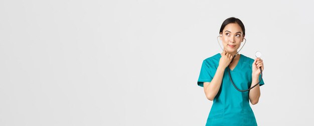 Covid-19, healthcare workers and preventing virus concept. Cute smiling asian female intern learn how listen lungs with stethscope, doctor examine patient, looking thoughtful upper left corner.