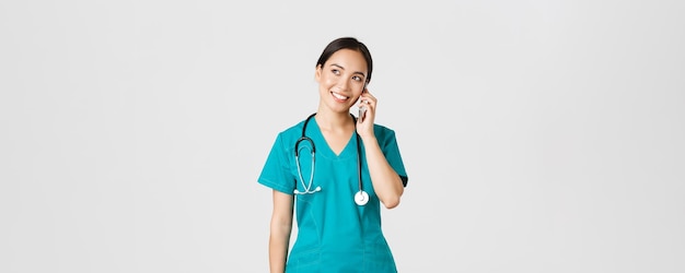 Covid-19, медицинские работники и концепция профилактики вирусов. Довольно улыбающаяся азиатская женщина-врач, врач в халате разговаривает, разговаривает по телефону и мечтательно смотрит в верхний левый угол
