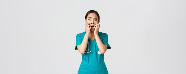 Covid-19, healthcare workers and preventing virus concept. Scared insecure asian female doctor, nurse in scrubs starts to panic from fear, biting fingernails and looking horrified, white background