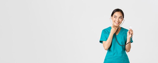 Covid-19, healthcare workers and preventing virus concept. Smiling cute asian doctor, female nurse examine patient lungs, using stethoscope, listening closer, standing white background.