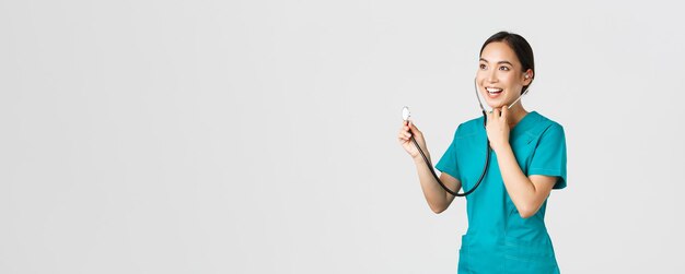 Covid-19, медицинские работники и концепция профилактики вирусов. Улыбающаяся дружелюбно выглядящая азиатская женщина-врач слушает легкие пациента во время ежедневного осмотра, используя стетоскоп, на белом фоне