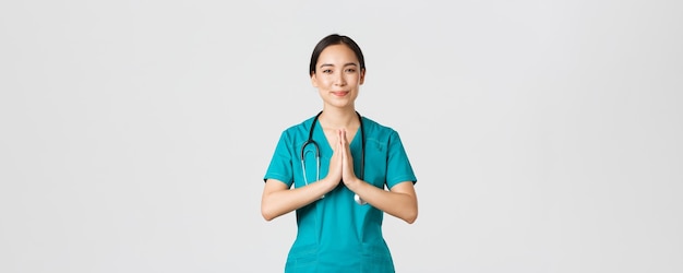 코비드-19, 의료 종사자 및 바이러스 개념 예방. 친절하고 돌보는 미소, 예쁜 아시아 의사, 수술복을 입은 여성 의사, 나마스테를 보여주는, 기도하는 몸짓, 흰색 배경