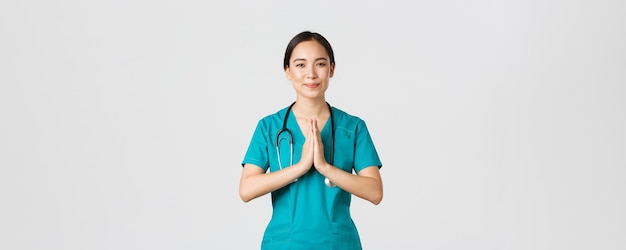 Covid-19, healthcare workers and preventing virus concept. Smiling kind and caring, pretty asian physician, female doctor in scrubs, showing namaste, praying gesture, white background