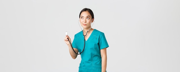 Covid-19, healthcare workers and preventing virus concept. Young focused asian female doctor, physician in scrubs, examine patient lungs, using stethoscope, standing thoughtful white background