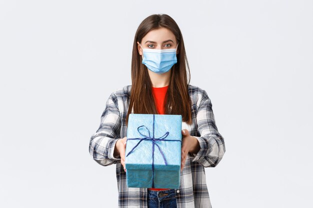 Covid-19, lifestyle, holidays and celebration concept. Cheerful young girl in medical mask stretch hand, handing birthday gift to you, smiling, congratulate with b-day, white background.