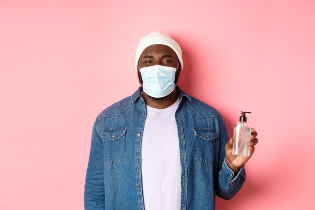 Covid-19, lifestyle and lockdown concept. Handsome hipster guy in face mask showing hand sanitizer, using antiseptic, standing over pink background