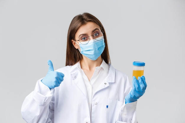 Covid-19, medical research, diagnosis, healthcare workers and quarantine concept. Doctor or clinic lab employee holding patient urine sample, wear medical mask and gloves, thumb-up