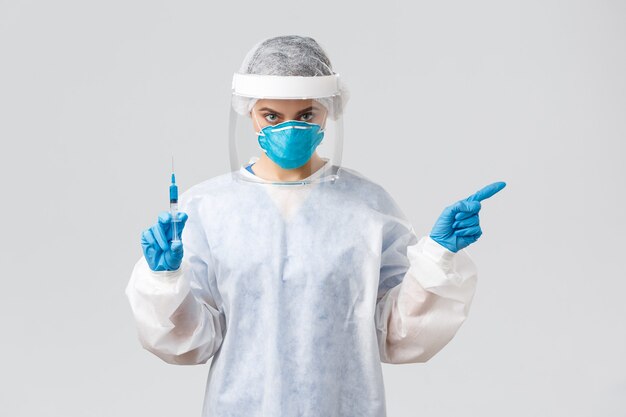 Covid-19, medical research, diagnosis, healthcare workers and quarantine concept. Serious doctor in personal protective costume, medical respirator, pointing left, hold syringe with vaccine