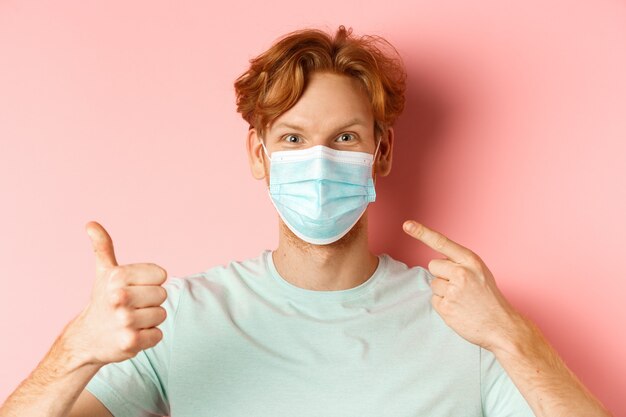 Covid-19 and pandemic concept. Handsome redhead guy pointing finger at face mask and showing thumbs-up, using measures from coronavirus, standing over pink background.