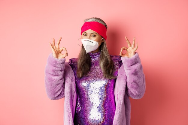 코비드-19, 전염병 및 패션 개념. 세련된 디스코 드레스와 인공 호흡기를 착용한 멋진 아시아 할머니, 괜찮은 표시를 보여주고, 얼굴 마스크와 사회적 거리, 분홍색 배경 착용을 요구합니다.