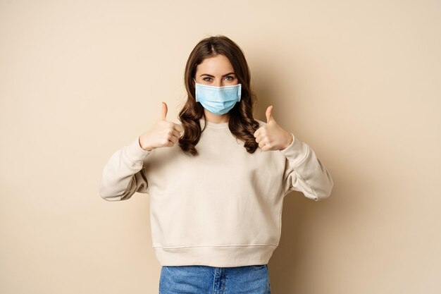 코비드-19, 전염병 및 검역 개념. 젊은 여성은 코로나바이러스 오미크론 발병 동안 의료용 안면 마스크를 쓰고 베이지색 배경 위에 엄지손가락을 보이고 서 있습니다.