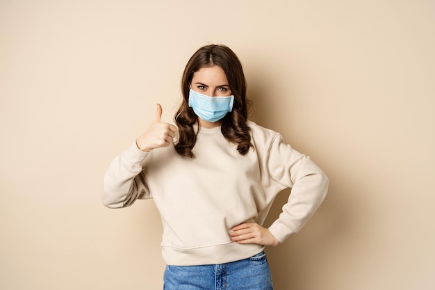Covid-19、パンデミックおよび検疫の概念。若い女性は、コロナウイルスオミクロンの発生時に医療用フェイスマスクを着用し、親指を立てて、ベージュの背景の上に立っています
