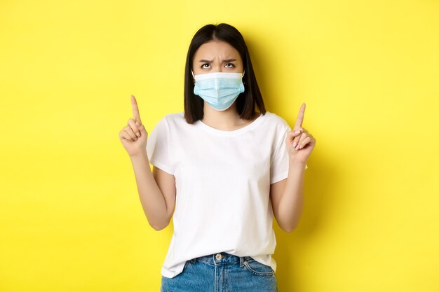 Covid-19, pandemic and social distancing concept. Disappointed asian girl in medical mask, frowning upset and pointing fingers up at logo, standing over yellow background