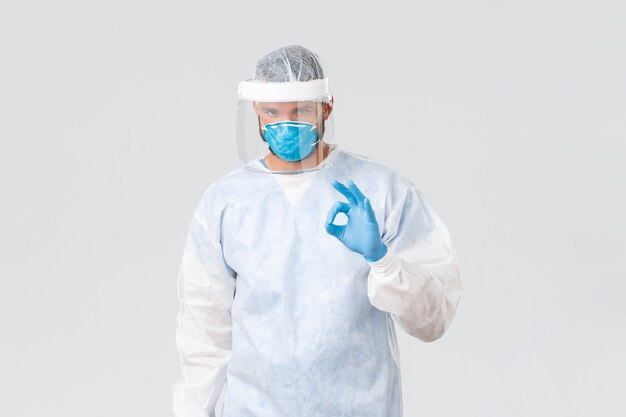 Covid-19 pandemic, virus outbreak, clinic and healthcare workers concept. Serious confident doctor in personal protective equipment, show okay sign, guarantee be on guard for patients.