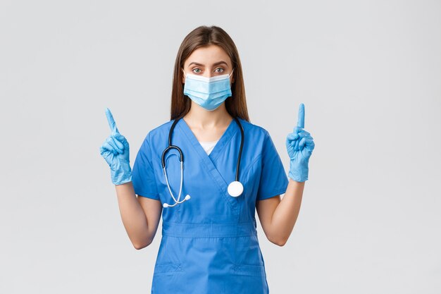 Covid-19, preventing virus, health, healthcare workers concept. Serious and confident female nurse in blue scrubs, medical mask PPE, pointing fingers up, inform patients how prevent corona infection