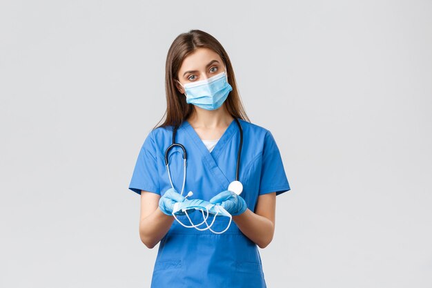Covid-19, preventing virus, health, healthcare workers and quarantine concept. Attractive female nurse in blue scrubs and personal protective equipment, giving medical masks to patient
