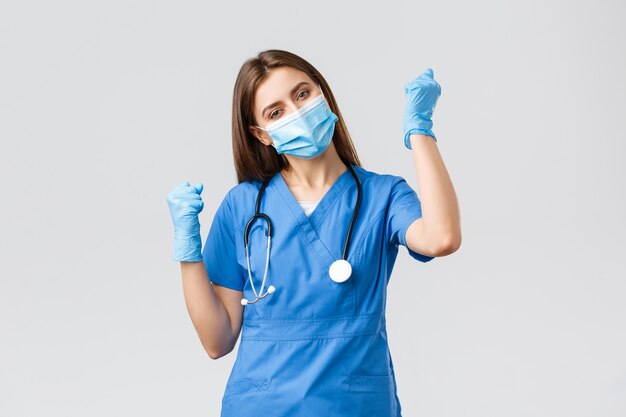 Covid-19、ウイルス、健康、医療従事者、検疫の概念を防ぎます。陽気な楽観的な女性看護師の勝利、勝利を祝う、明るい拳ポンプ、医療用マスクと手袋で笑う